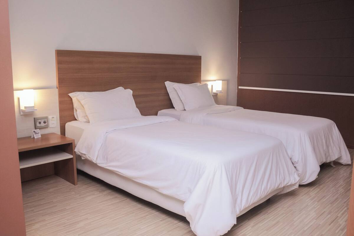 A hotel room with two beds, wooden headboards, white bedding, bedside tables, and wall-mounted lamps.