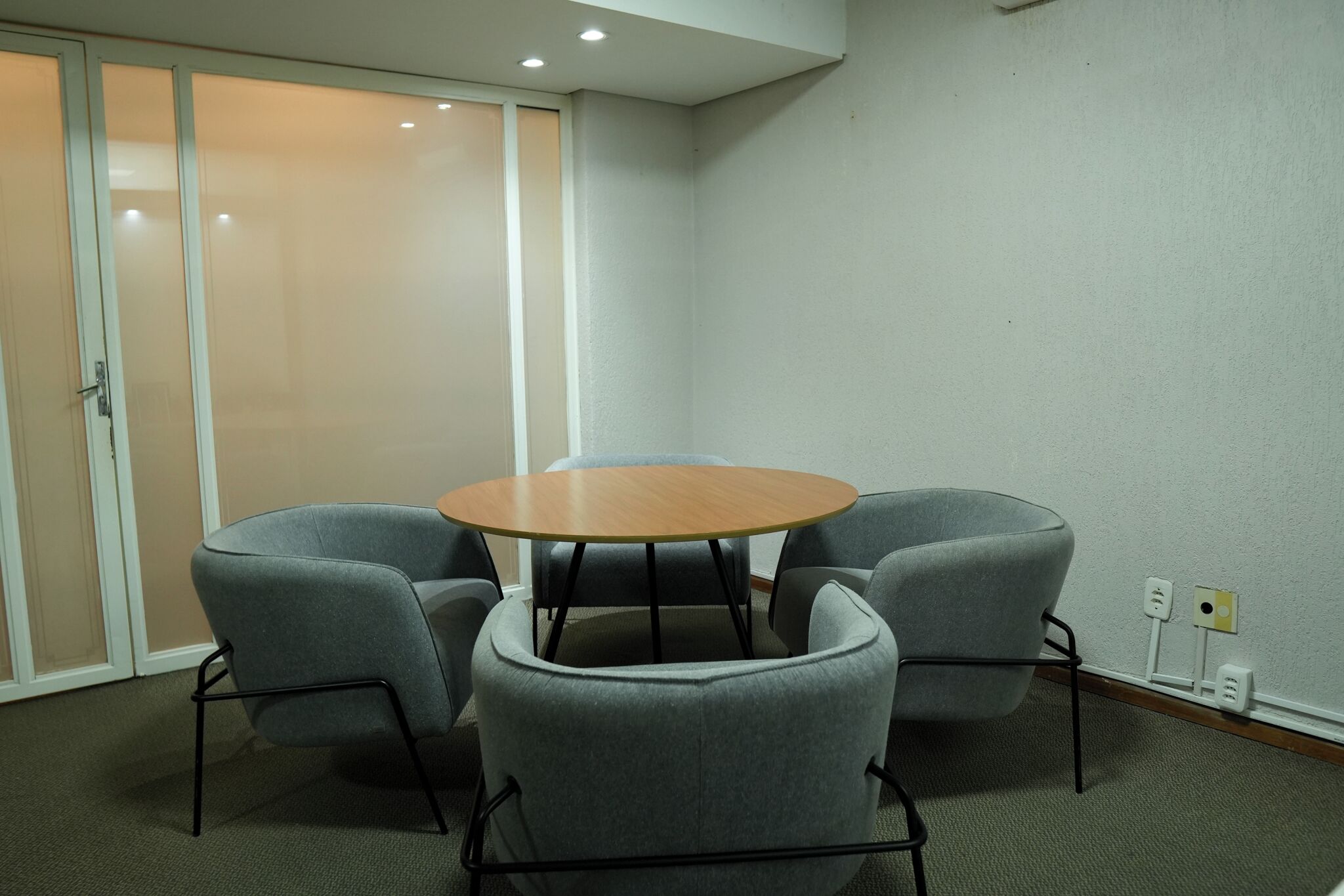 Una sala de conferencias con una mesa redonda y cuatro sillas grises, una puerta corredera de vidrio y un enchufe de pared.