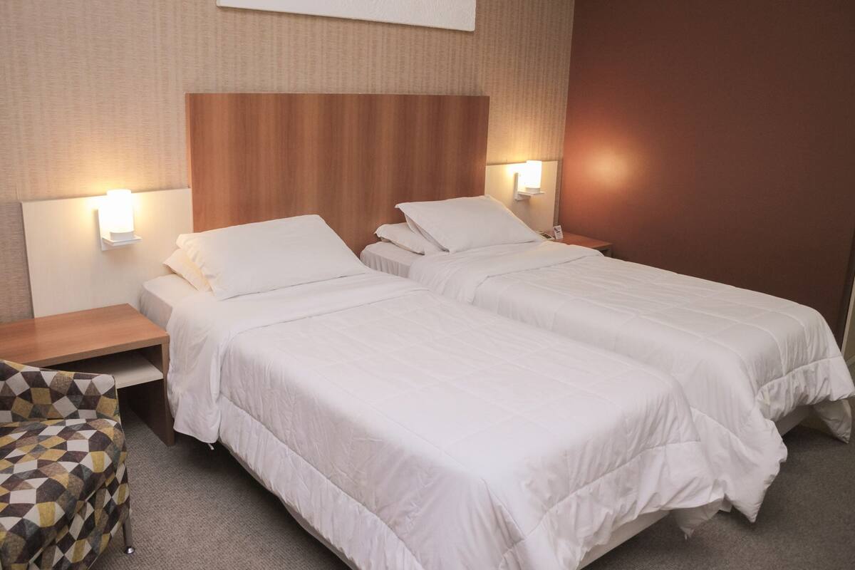 A hotel room with two beds, a wooden headboard, bedside lamps, and a patterned armchair.