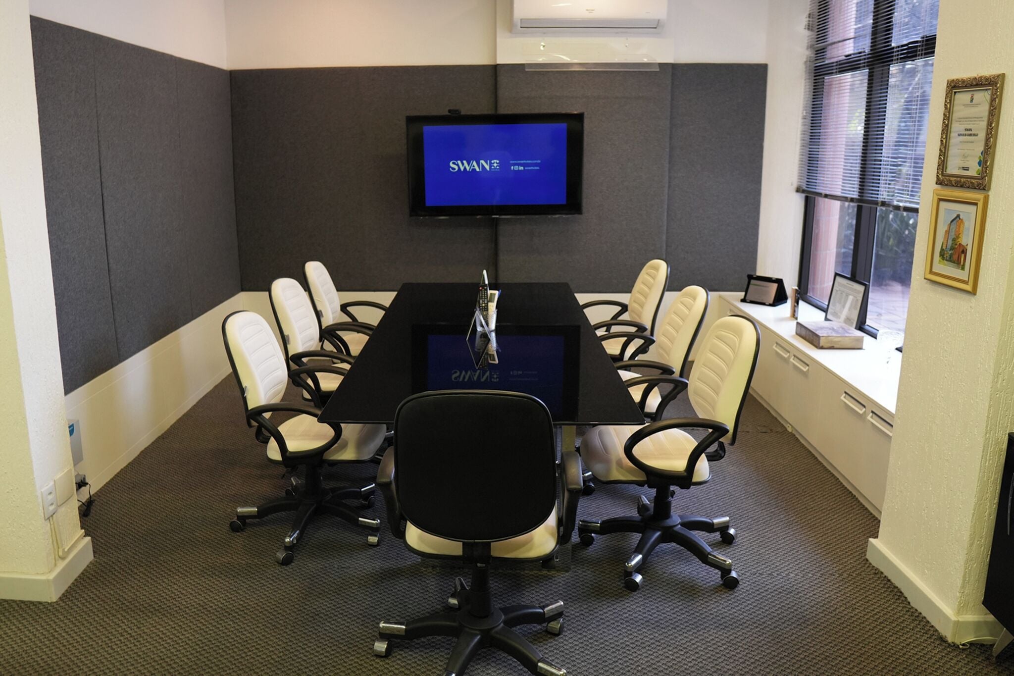 Un salón de conferencias con una mesa larga, seis sillas, una pantalla de televisión y un ventanal con persianas.
