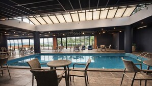 Una piscina interior con sillas de descanso y mesas, ventanas amplias y un diseño moderno en el techo.