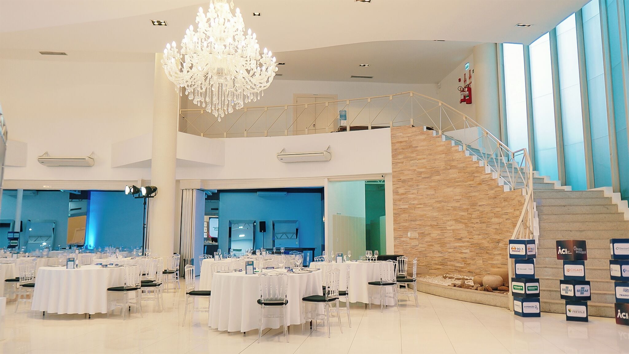 A spacious hall with multiple round tables, a grand chandelier, a staircase, and a modern staircase with glass railings.