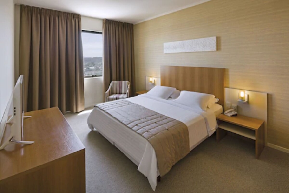 Habitación de hotel con una cama grande, un escritorio con computadora, una silla, una mesita de noche con lámpara y vistas a la ciudad desde la ventana.
