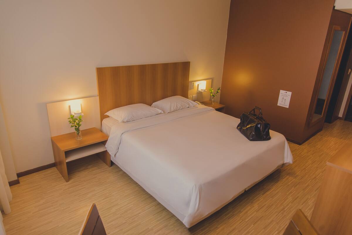 A hotel room with a wooden headboard, two bedside tables with lamps, a bed with white linens, and a handbag on the bed.