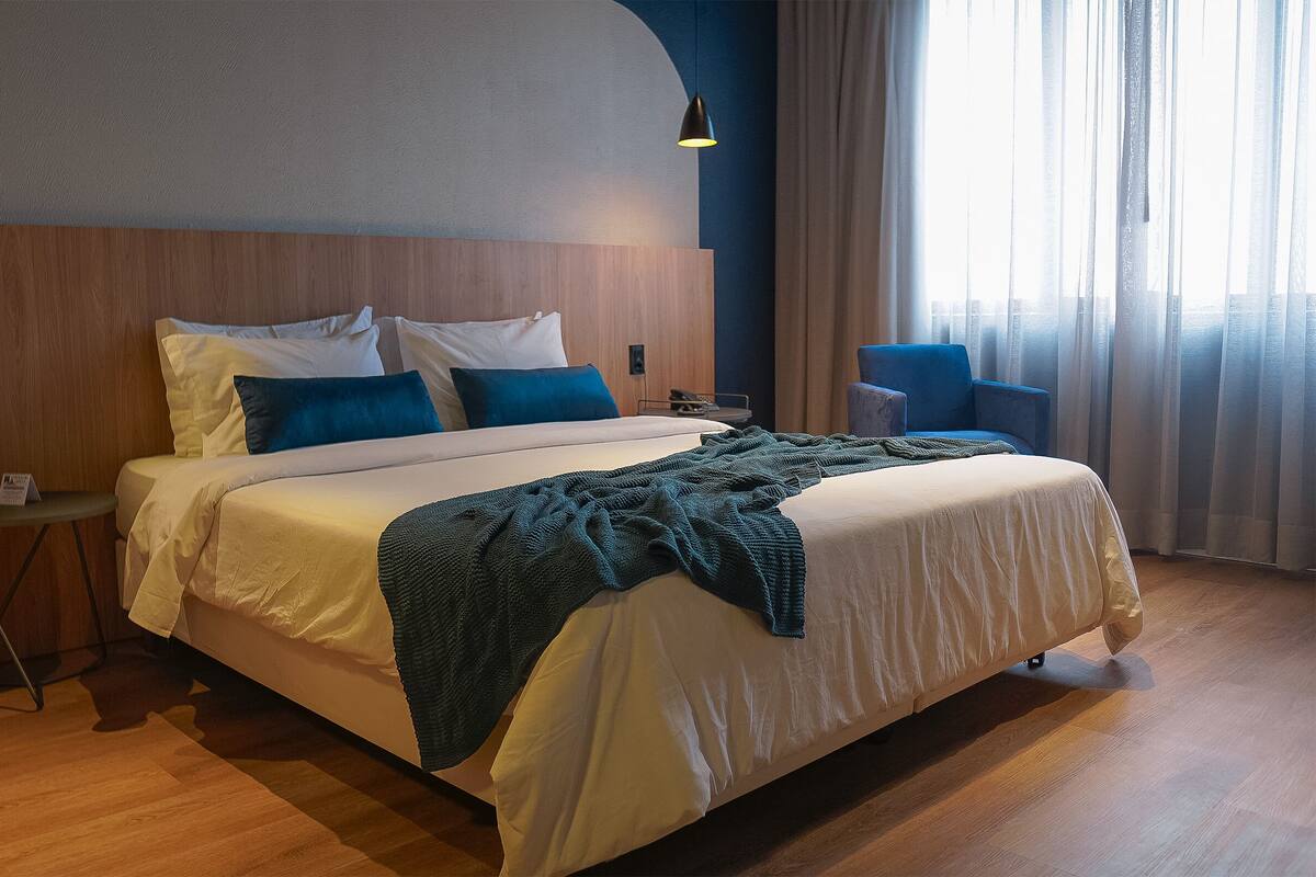 A neatly made bed with a blue blanket, a bedside table with a lamp, and a chair by the window.