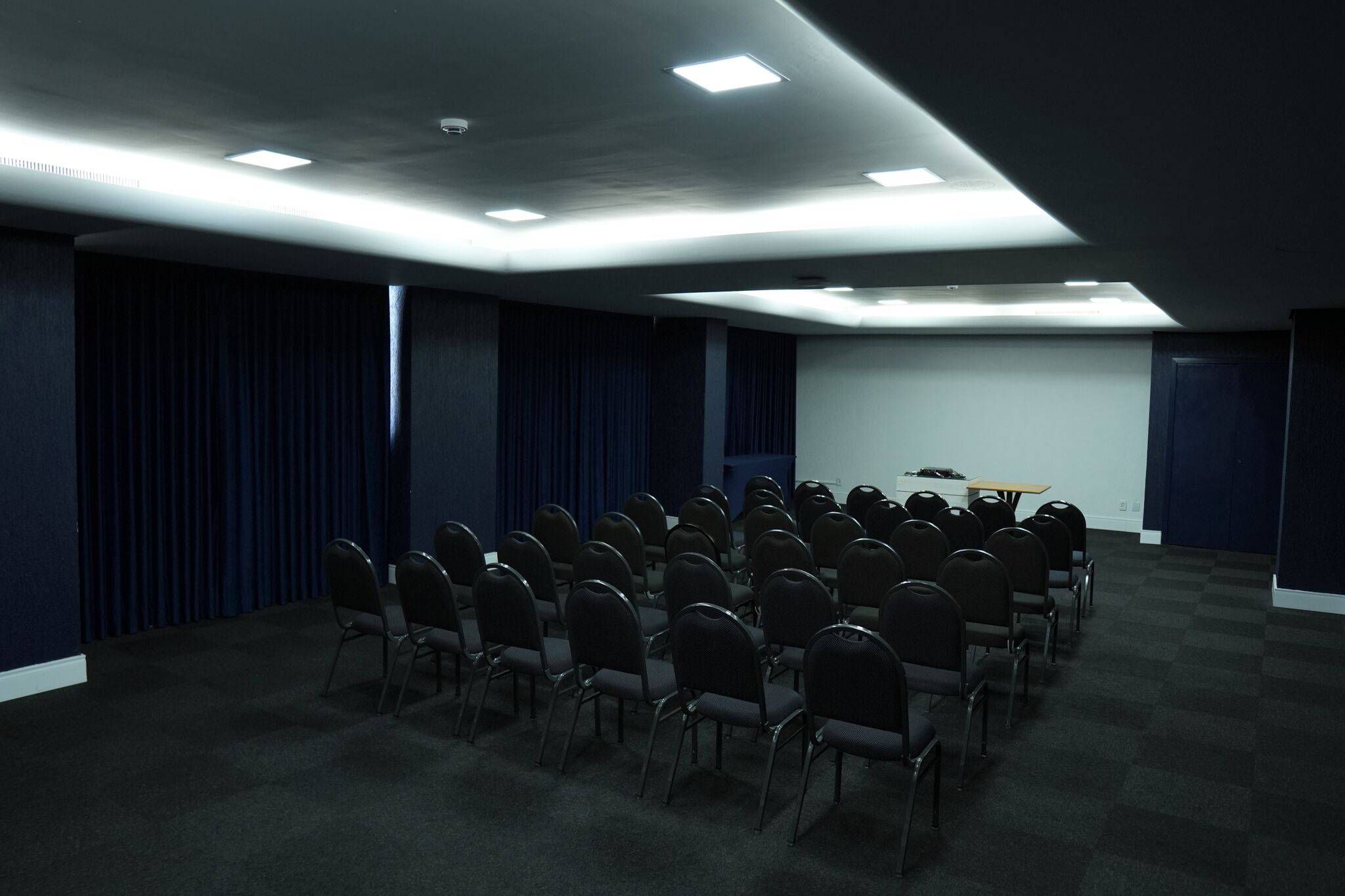 Un salón de conferencias con filas de sillas, una pantalla de proyector y una mesa con una computadora portátil.
