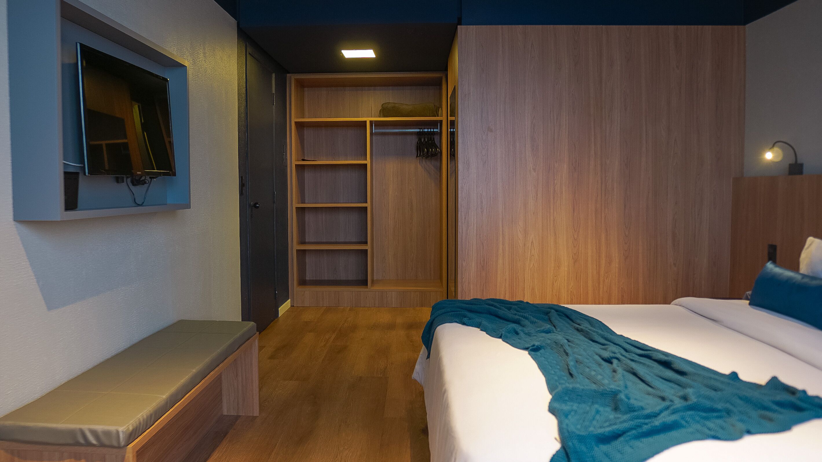 A hotel room with a bed, a bench, a TV mounted on the wall, a wardrobe, and a closet.