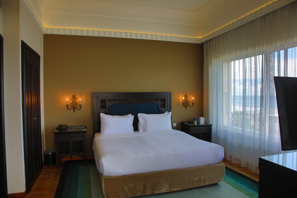 A hotel room with a large bed, two bedside tables, a TV, and a view of the city through sheer curtains.
