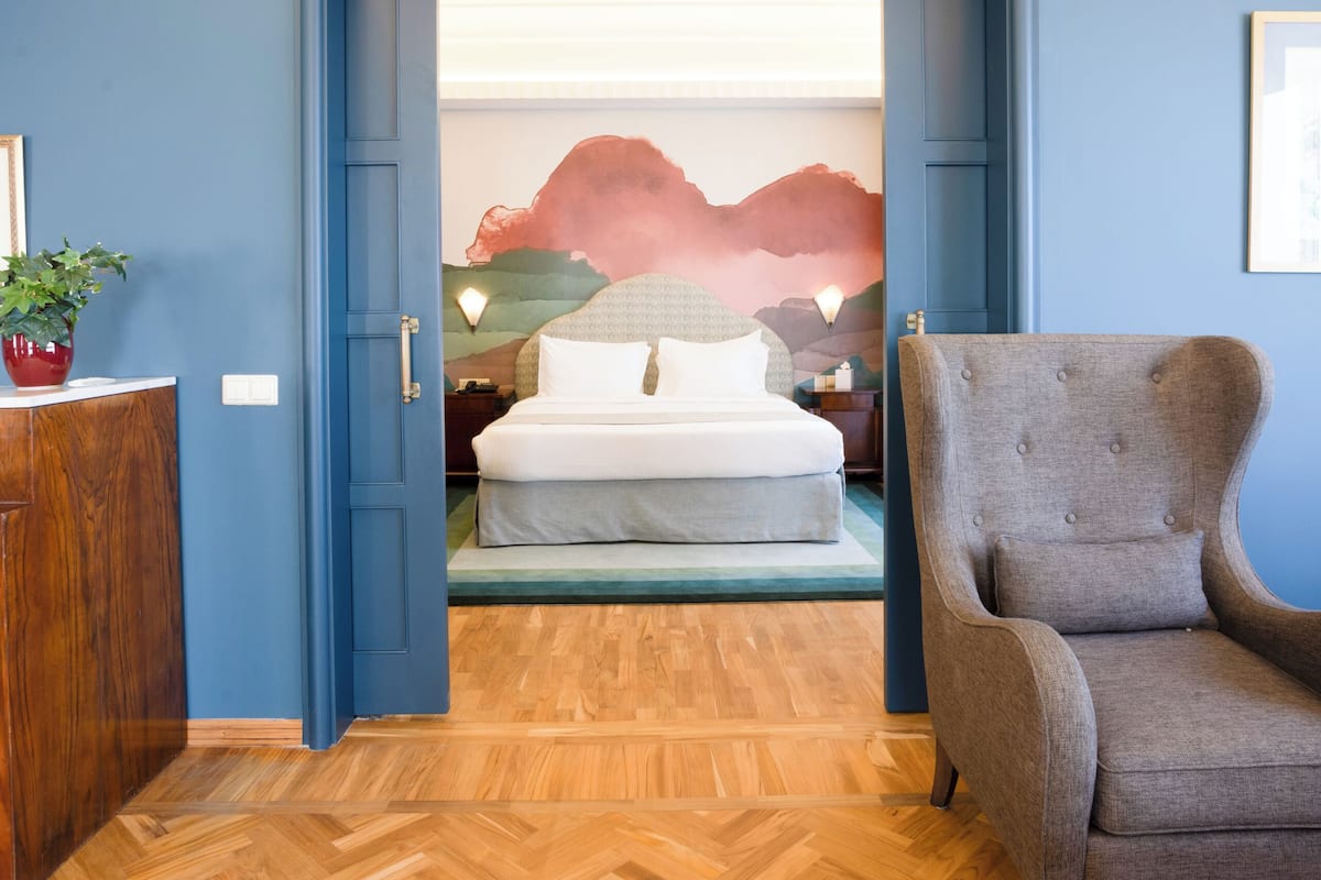 A room with a wooden herringbone floor, a grey armchair, a bed with white bedding, and a wall with a painting.