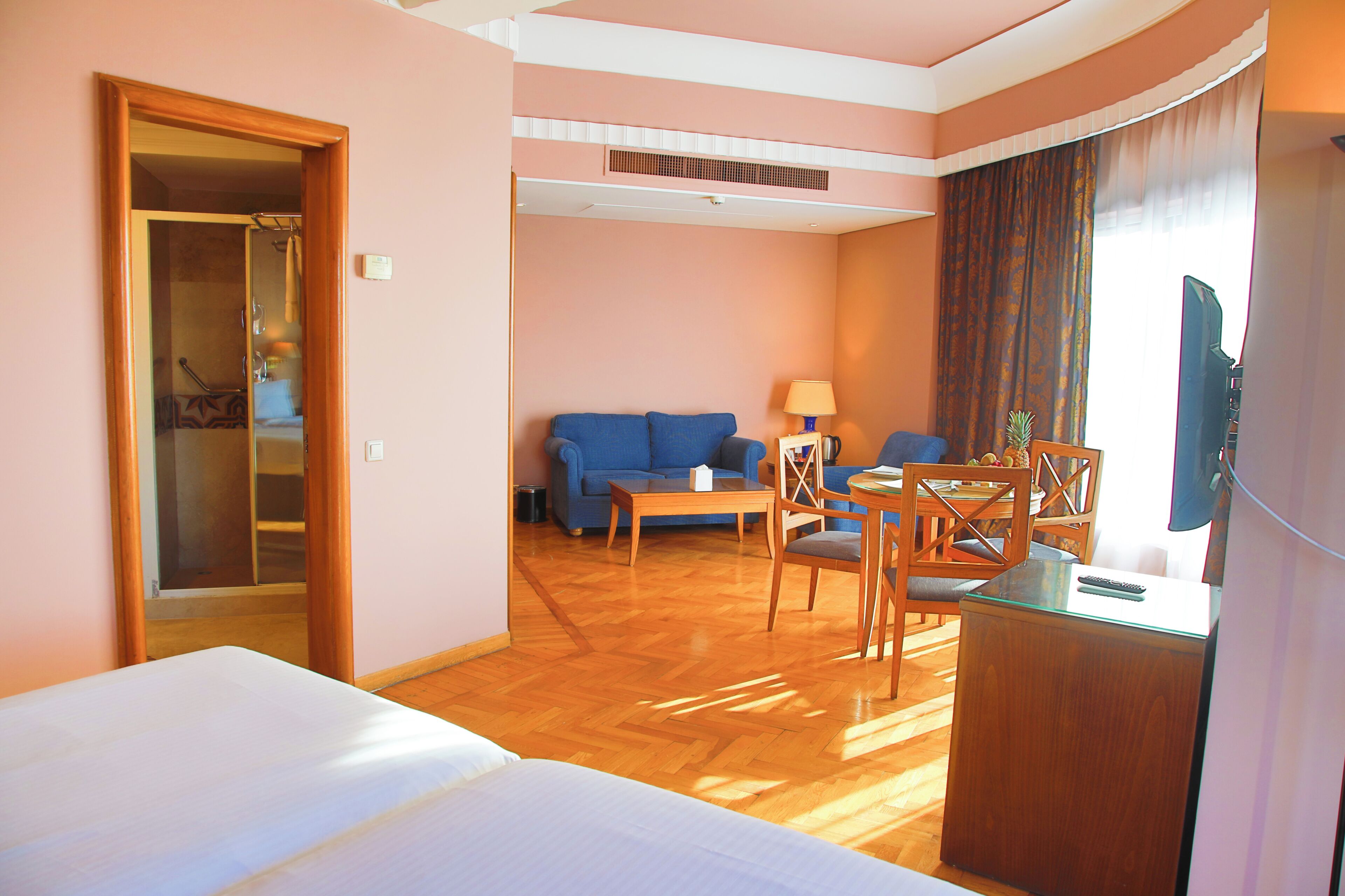 Habitación de hotel con cama, sofá, mesa de comedor con sillas, televisión y baño visible a través de una puerta.