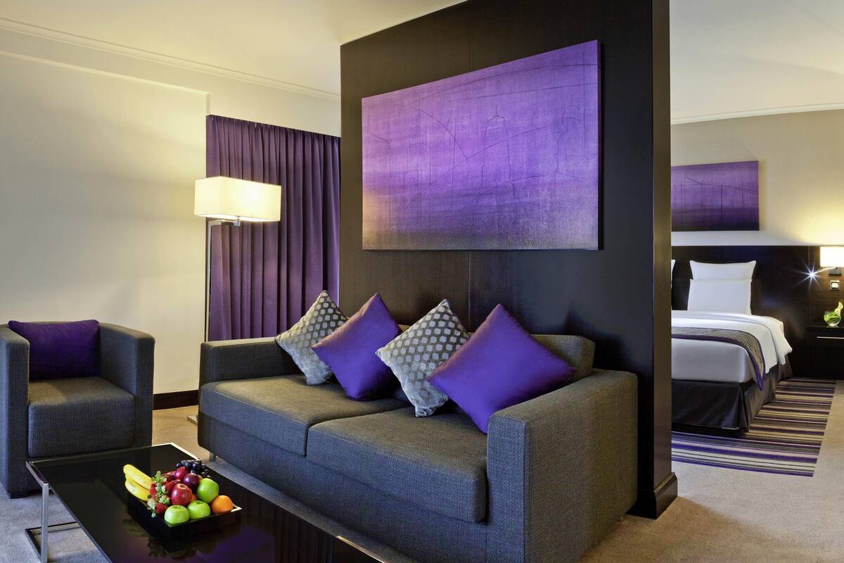 A modern hotel room with a grey sofa, purple cushions, a black coffee table with fruit, and a large purple abstract painting.