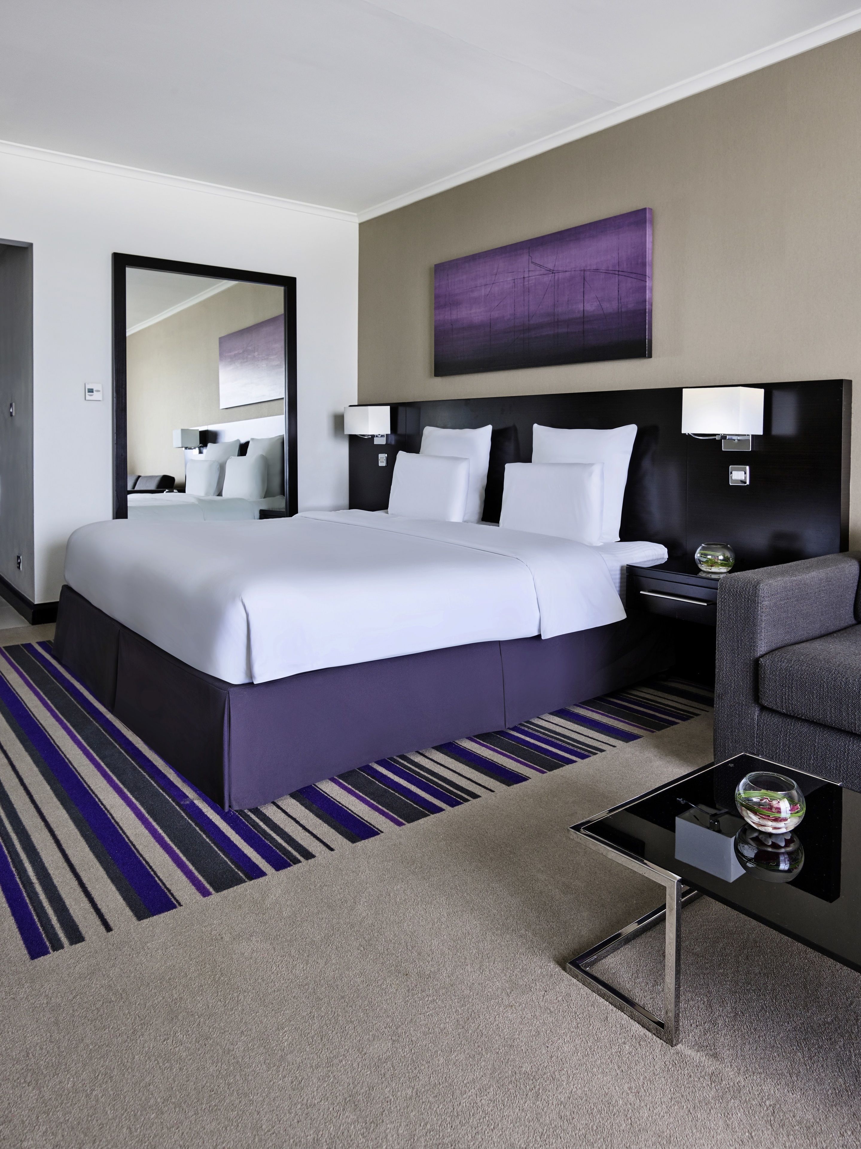 Une chambre d’hôtel moderne avec un grand lit, une table de chevet avec une lampe, une chaise et une petite table ornée d’un objet décoratif.