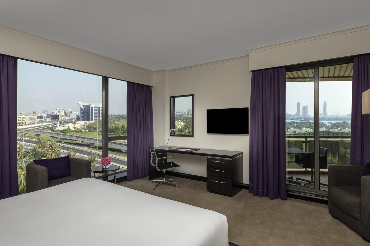 A hotel room with a large bed, a desk, a chair, a TV, and a balcony with a city view.