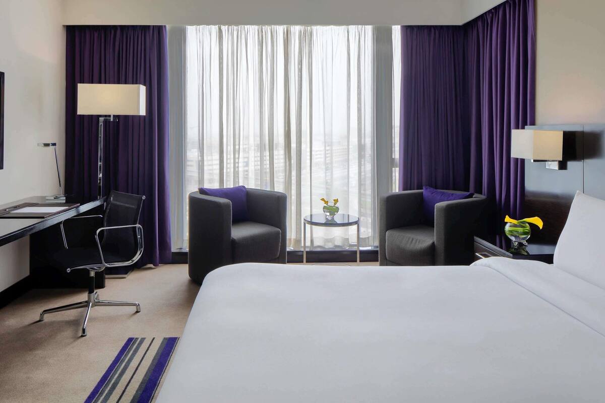 A hotel room with a bed, two armchairs, a desk, a lamp, a small table with a vase, and a view through large windows.
