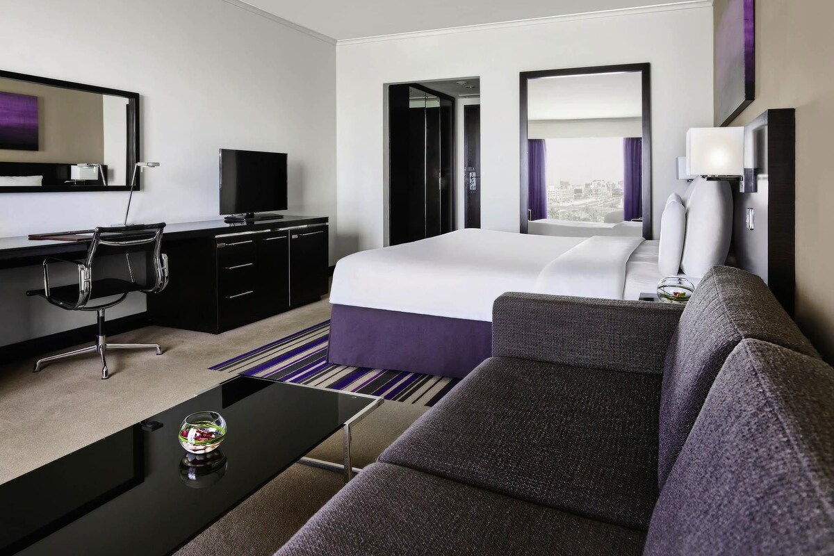 A modern hotel room with a bed, a desk with a TV, a sofa, and a large window.