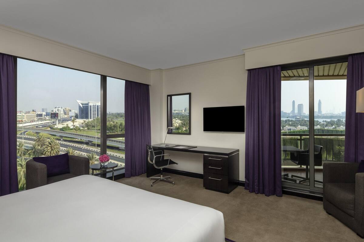 A hotel room with a large bed, a desk, a chair, a TV, and a balcony with a city view.