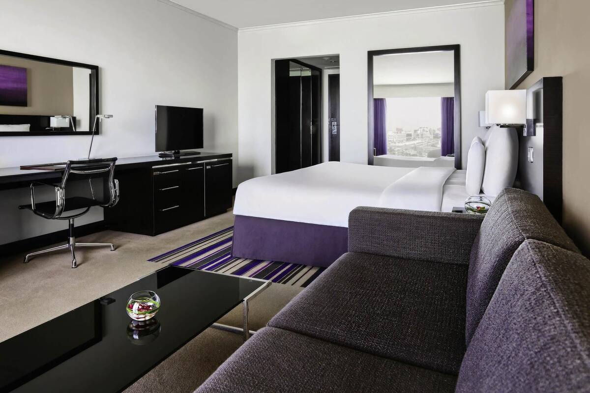 Una camera d'albergo moderna con un letto, una scrivania con televisore, un divano e un'ampia finestra.