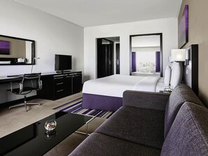 Quarto de hotel com cama, escrivaninha e cadeira, televisão, sofá e janela com cortinas.