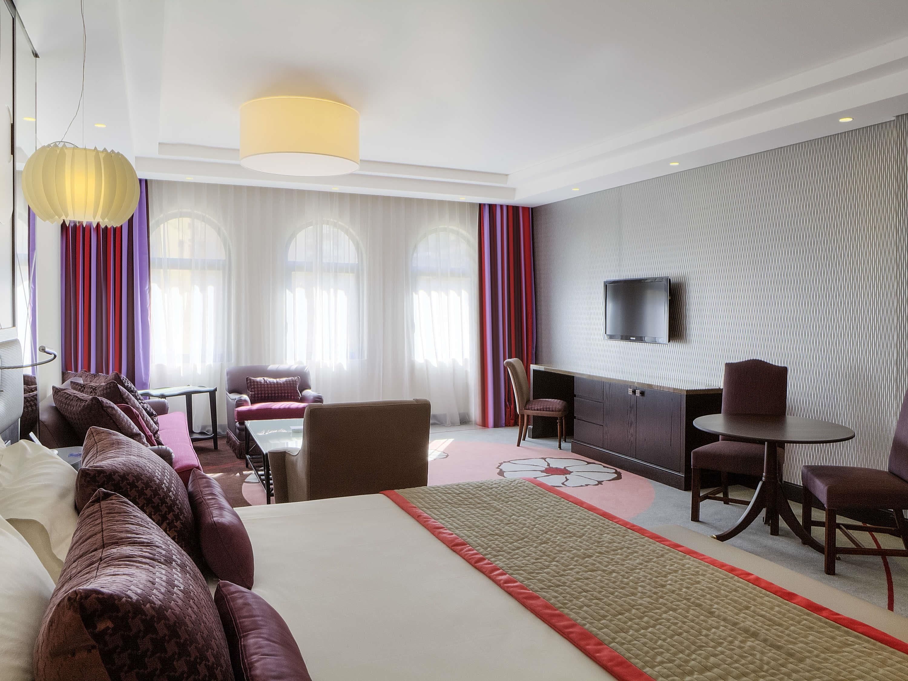 A hotel room with a large bed, a desk, a TV, and a seating area with chairs and a sofa.