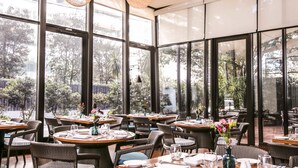 Un restaurant avec des tables rondes et des chaises en osier, de grandes fenêtres et des luminaires suspendus.