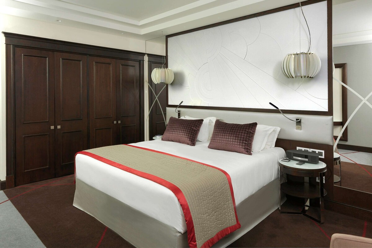 Habitación de hotel con una cama grande, un escritorio, una silla, un armario y un cuadro abstracto de grandes dimensiones en la pared.