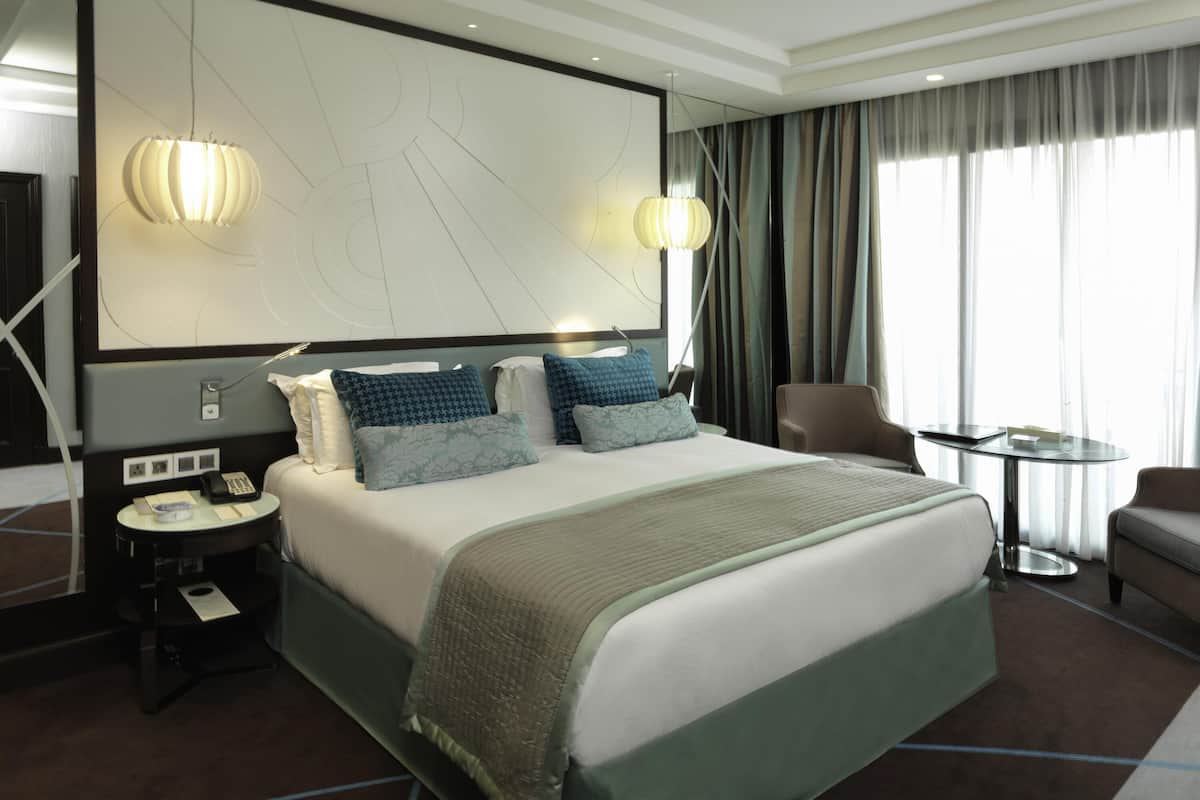 Habitación de hotel con una cama grande, mesitas de noche, un escritorio, una silla, un espejo y vistas al exterior.