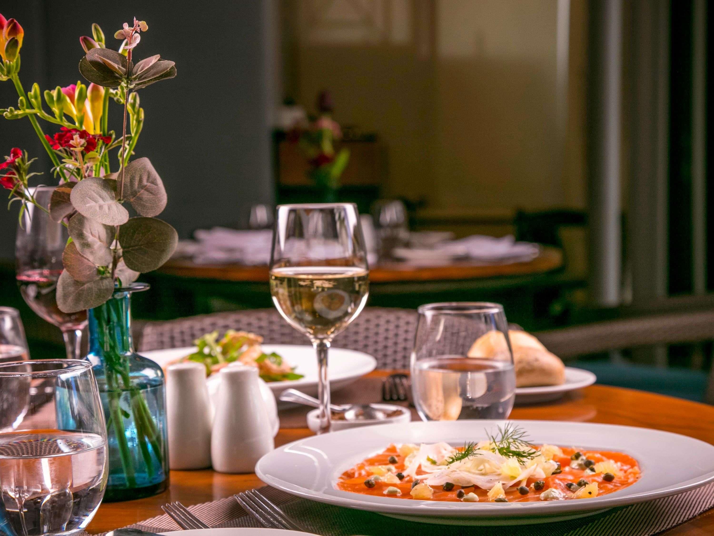 Une table à manger avec une assiette de nourriture, un verre de vin blanc et un vase de fleurs.