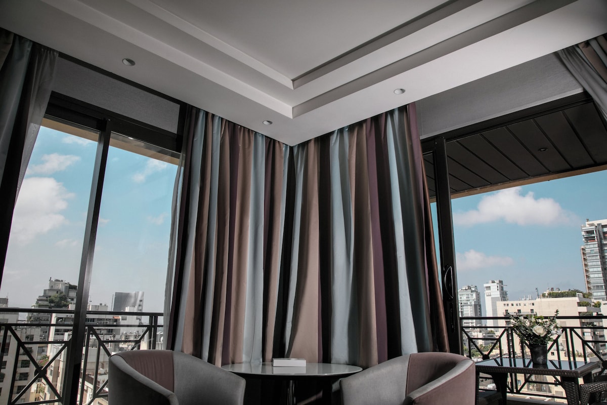 Una habitación moderna con una mesa redonda y dos sillas, ventanas amplias con cortinas y vistas a la ciudad.