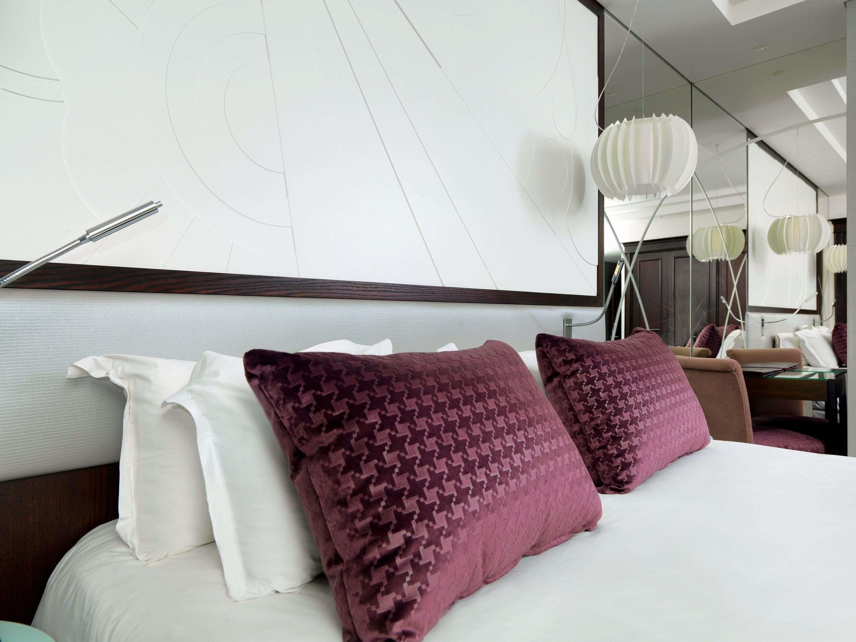 A hotel room with a large bed, two patterned pillows, a bedside table, a mirror, and a wall with a geometric design.