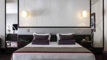 A modern hotel room with a large bed, two bedside lamps, a headboard with a geometric design, and a small bedside table with a cup and books.