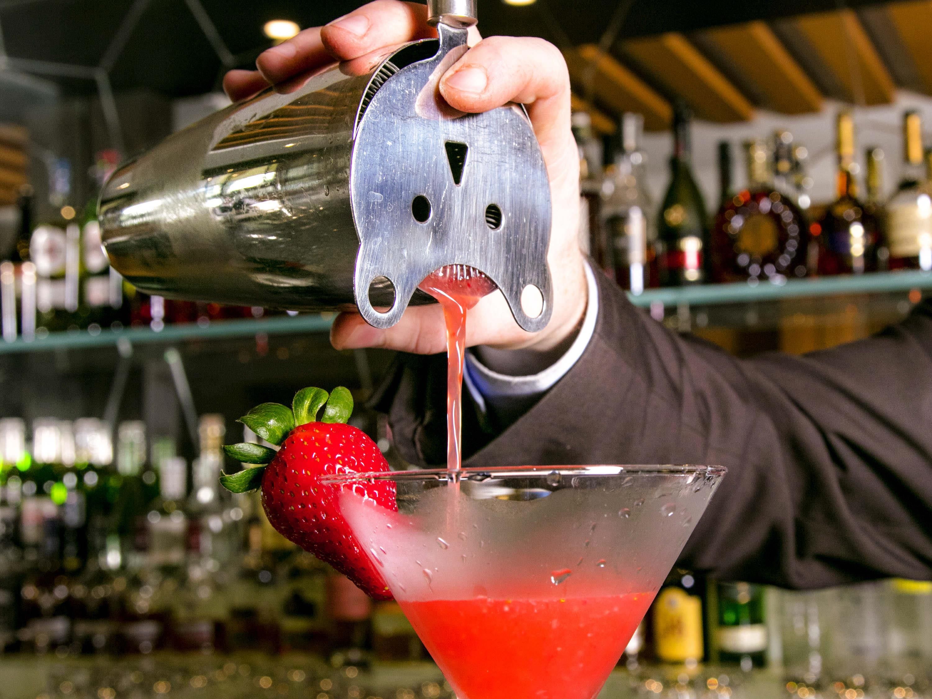 A bartender pouring a red cocktail into a martini glass with a strawberry garnish.