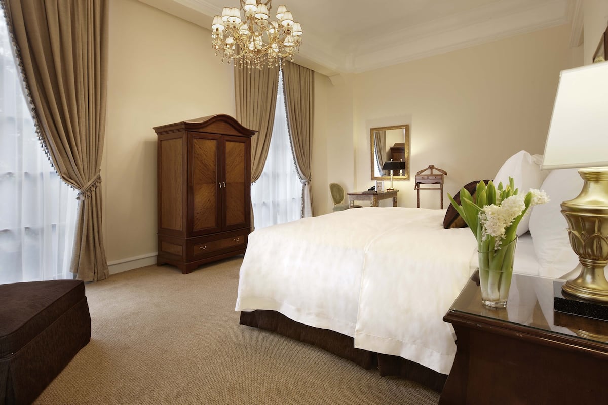 A hotel room with a large bed, a wooden wardrobe, a chandelier, a bedside table with a lamp, and a vase with flowers.