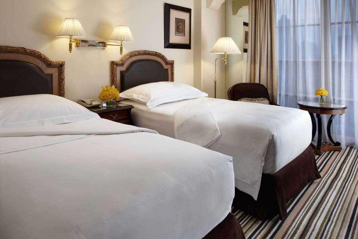 A hotel room with two beds, a nightstand with a lamp, a small table with a vase of flowers, and a window with sheer curtains.
