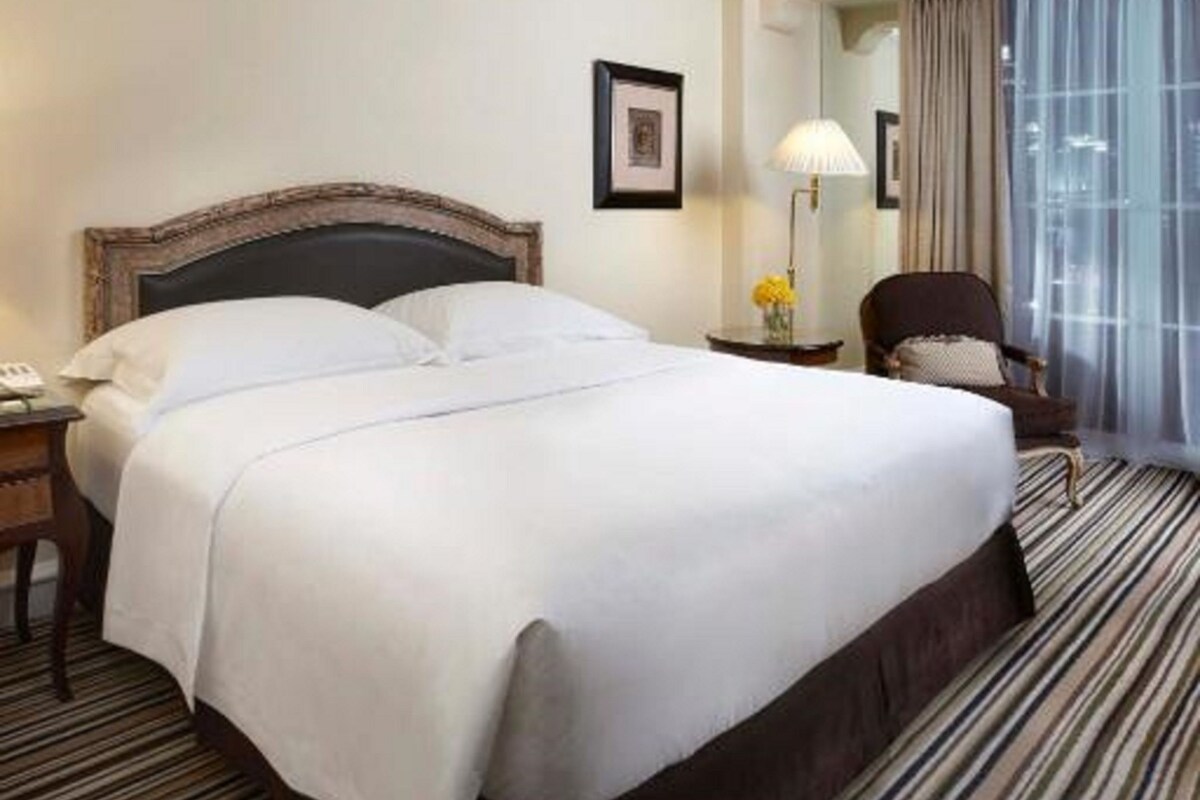 A hotel room with a large bed, a nightstand, a chair, a lamp, and a window with curtains.