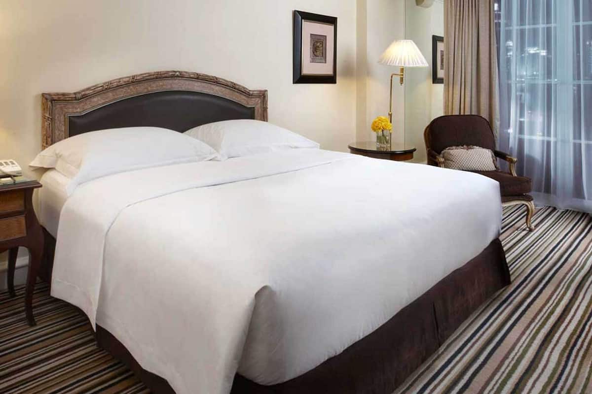 A hotel room with a large bed, a bedside table with a lamp, a chair, and a view of the city through the window.