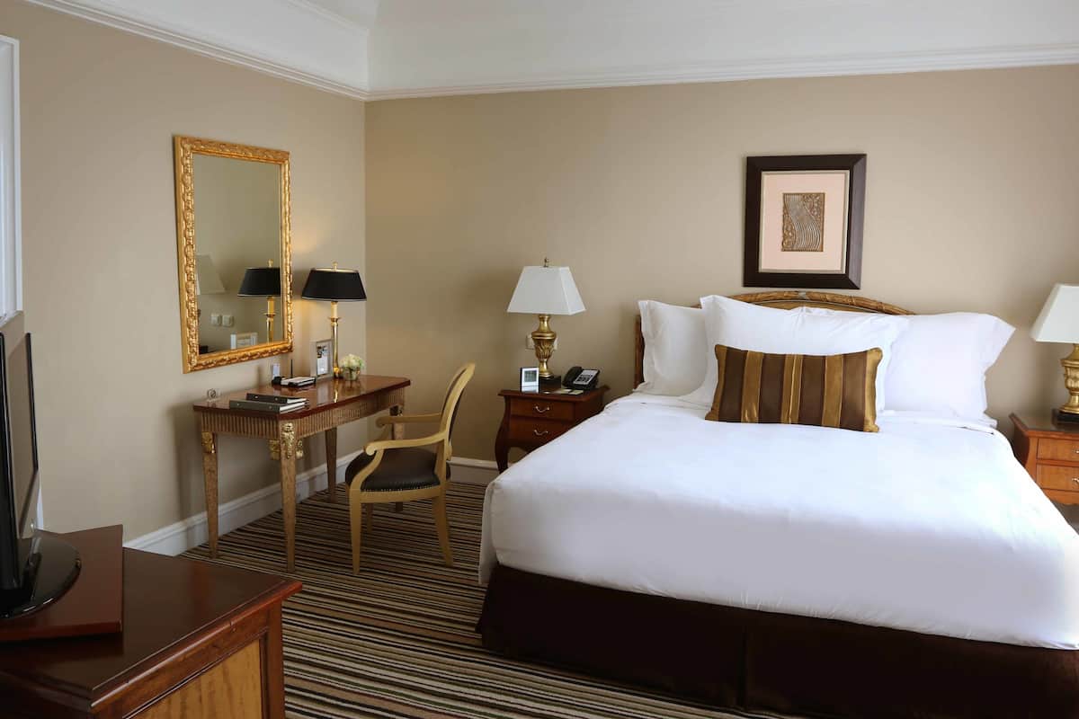 A hotel room with a large bed, a desk with a chair, a desk lamp, a telephone, and a framed picture on the wall.