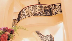 Een statige spiraaltrap met gedetailleerde zwarte leuningen, een grote bloemenvaas, en een luxe interieur met een vleugelpiano en elegante meubels.