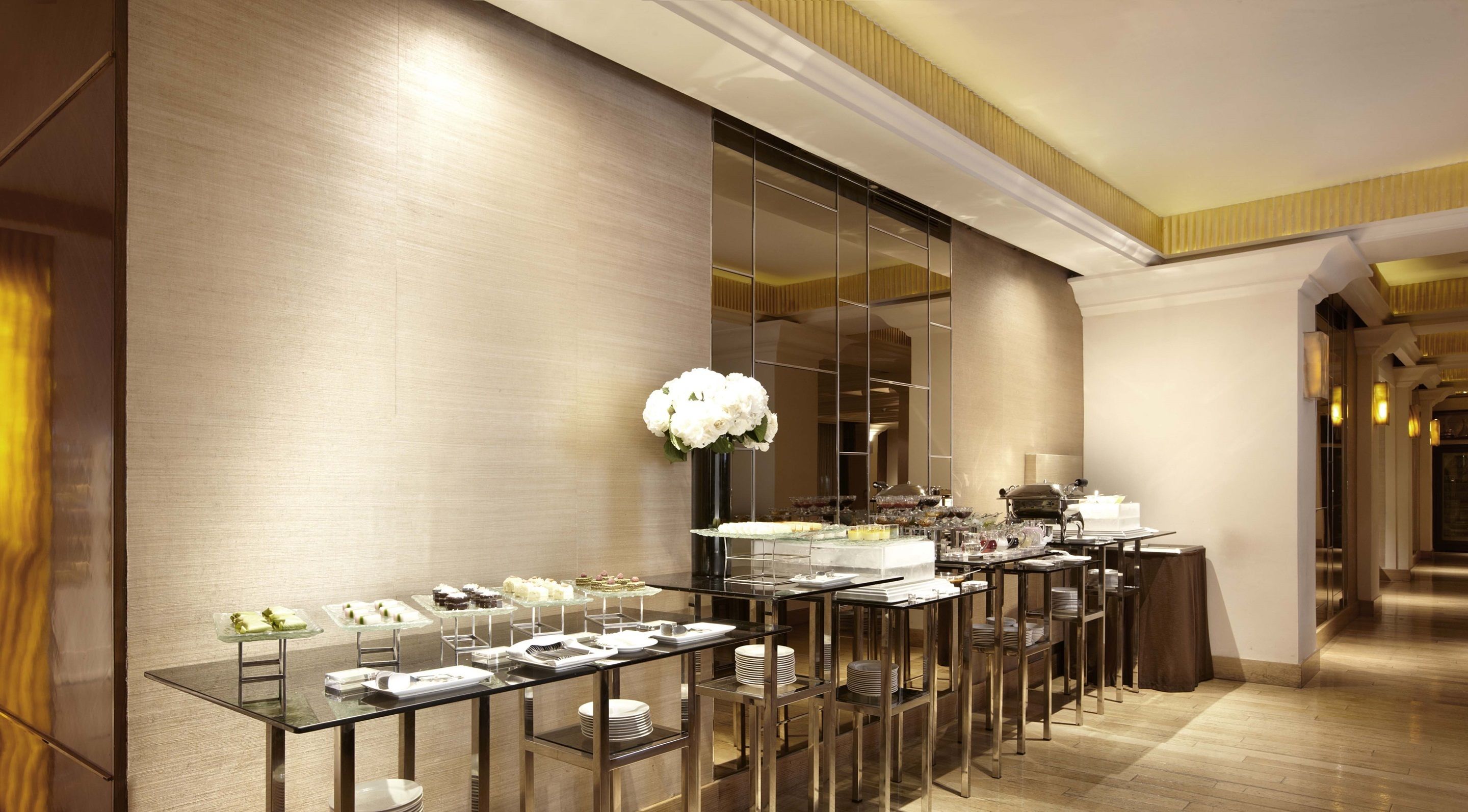 Un comedor de hotel moderno con mesas de vidrio y sillas de metal, un espejo grande y un centro de mesa floral blanco.