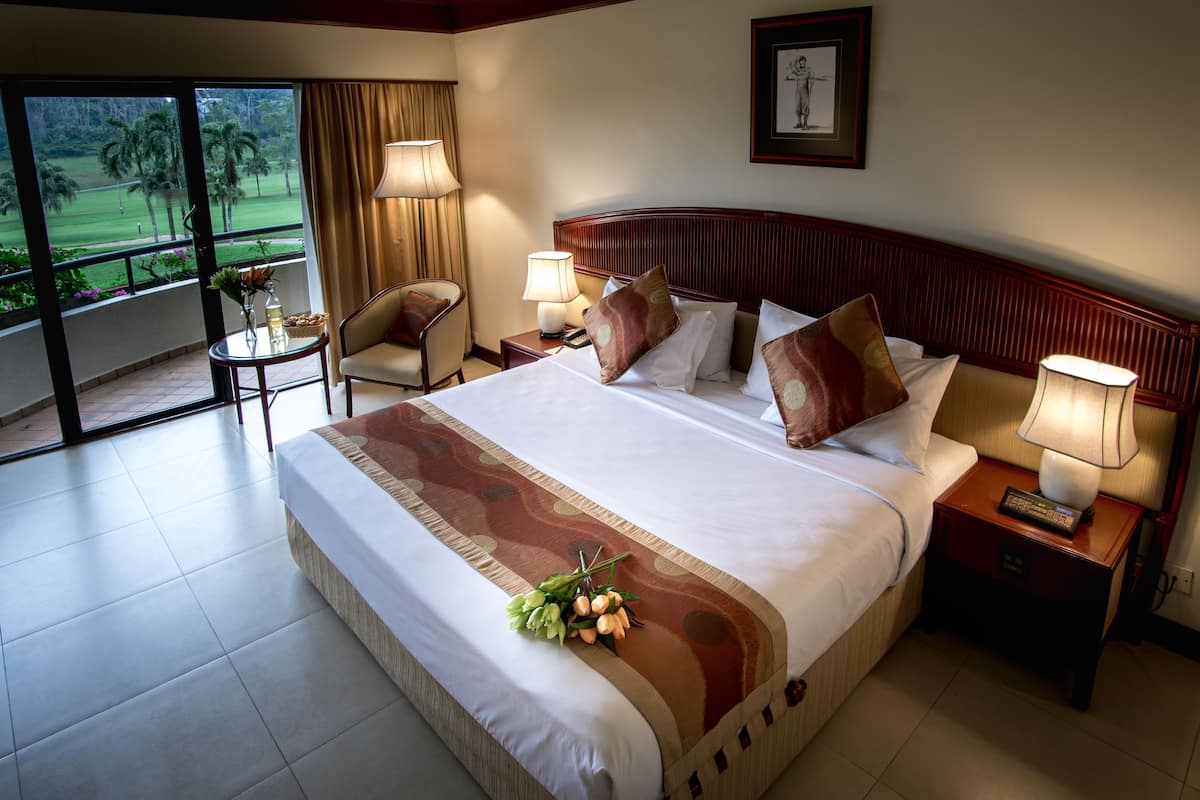 A hotel room with a large bed, two bedside tables with lamps, a chair, a small table with flowers, and a view of a golf course through the window.