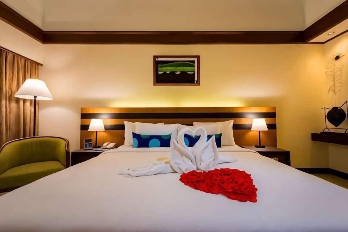 A hotel room with a bed, pillows, a nightstand, a lamp, a chair, and a framed picture on the wall.