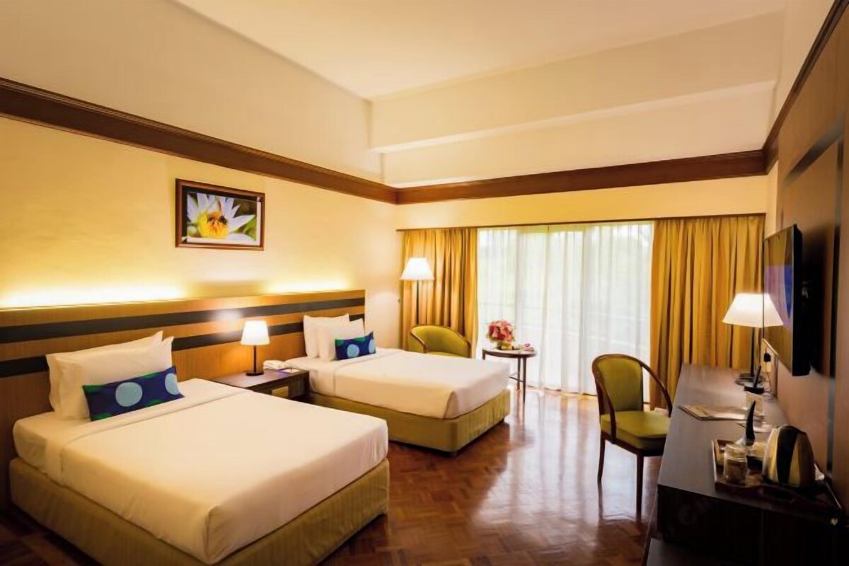 Quarto de hotel com duas camas, uma escrivaninha, uma cadeira, uma mesinha de cabeceira com abajur e uma janela com cortinas.