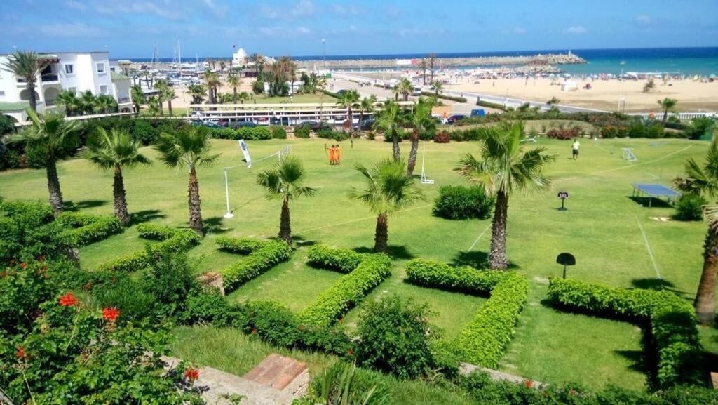 Un jardin bien entretenu, agrémenté de palmiers et de haies, avec une vue sur la plage et l’océan, et un bâtiment doté d’un balcon en arrière-plan.