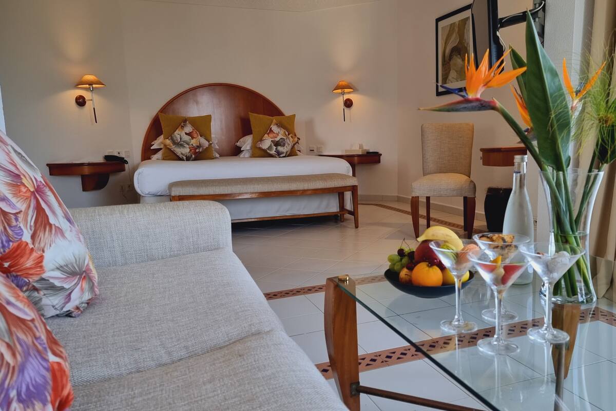 A hotel room with a bed, a sofa, a dining table with fruit and wine glasses, and a decorative plant.