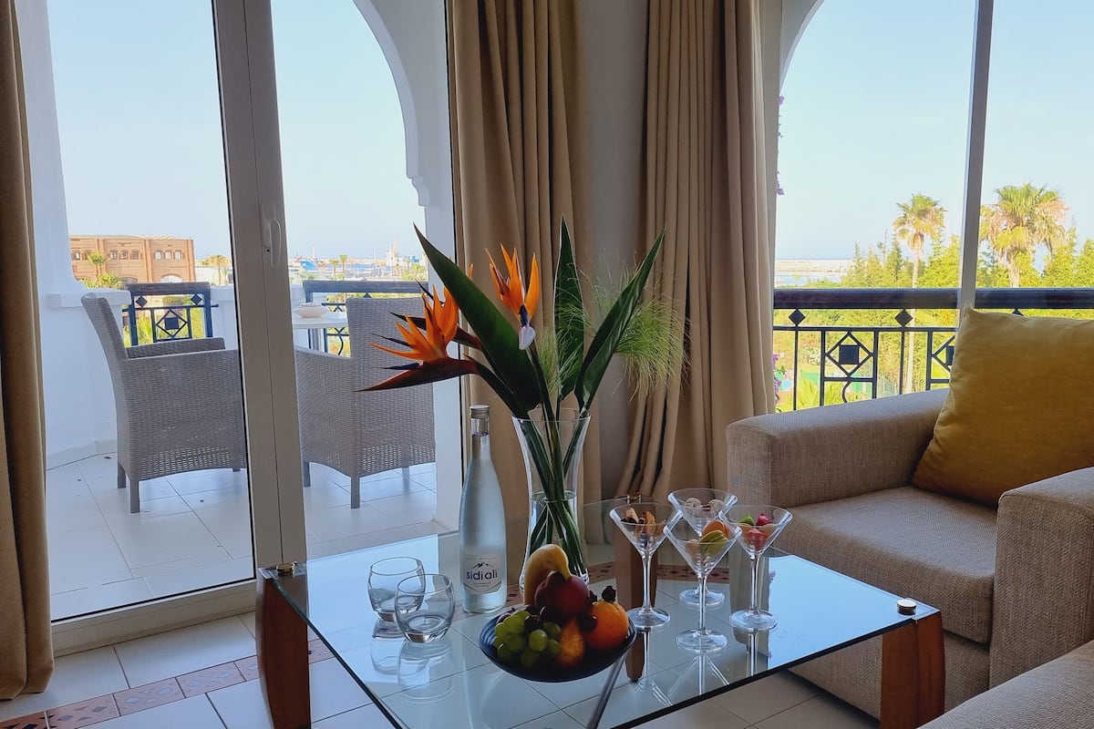 A modern living room with a glass coffee table, a sofa with a yellow pillow, and a balcony with wicker chairs and a view of palm trees.