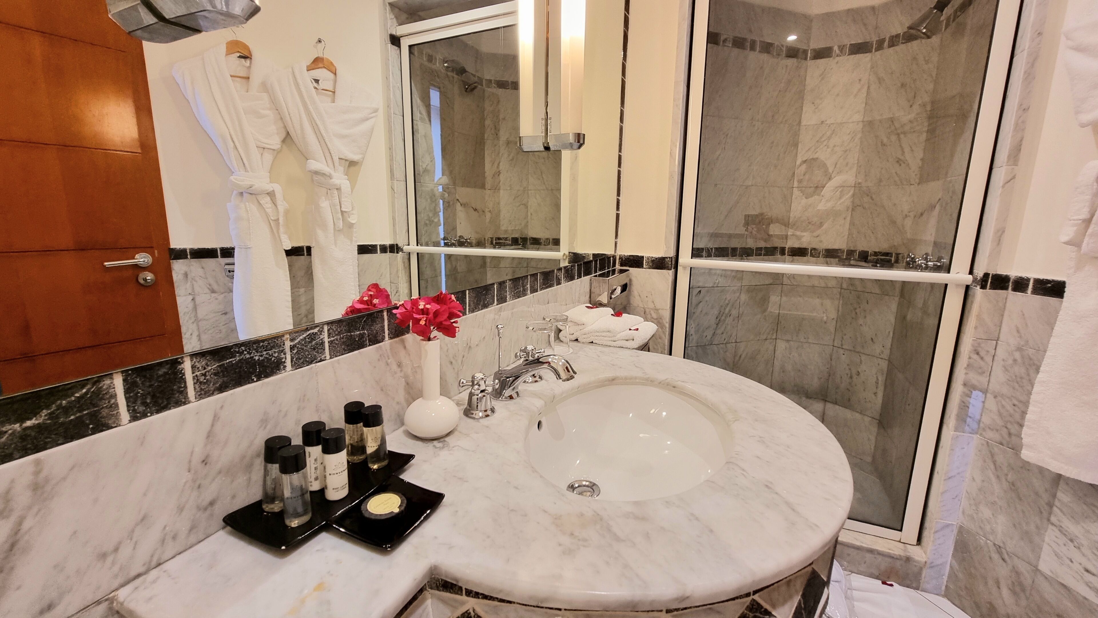 A bathroom with a marble sink, a glass shower enclosure, and a mirror reflecting a robe and a flower.