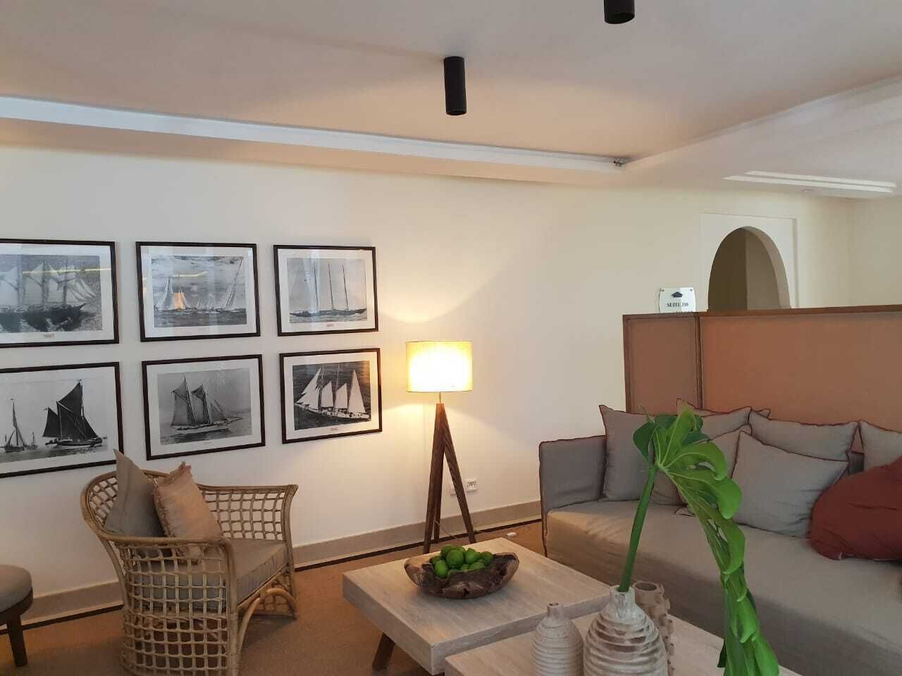 Un salon avec un canapé, un fauteuil en osier, une table basse sur laquelle se trouve un panier de fruits, une lampe et des tableaux encadrés accrochés au mur.