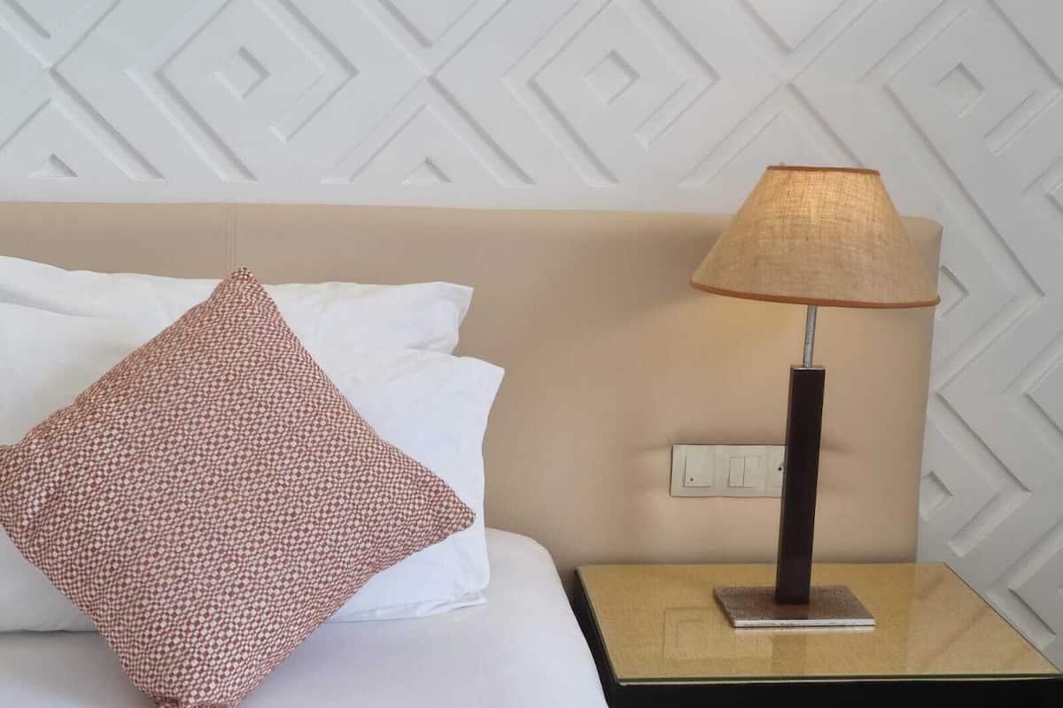 A neatly made bed with white and patterned pillows, a lamp on a bedside table, and a geometric wall design.