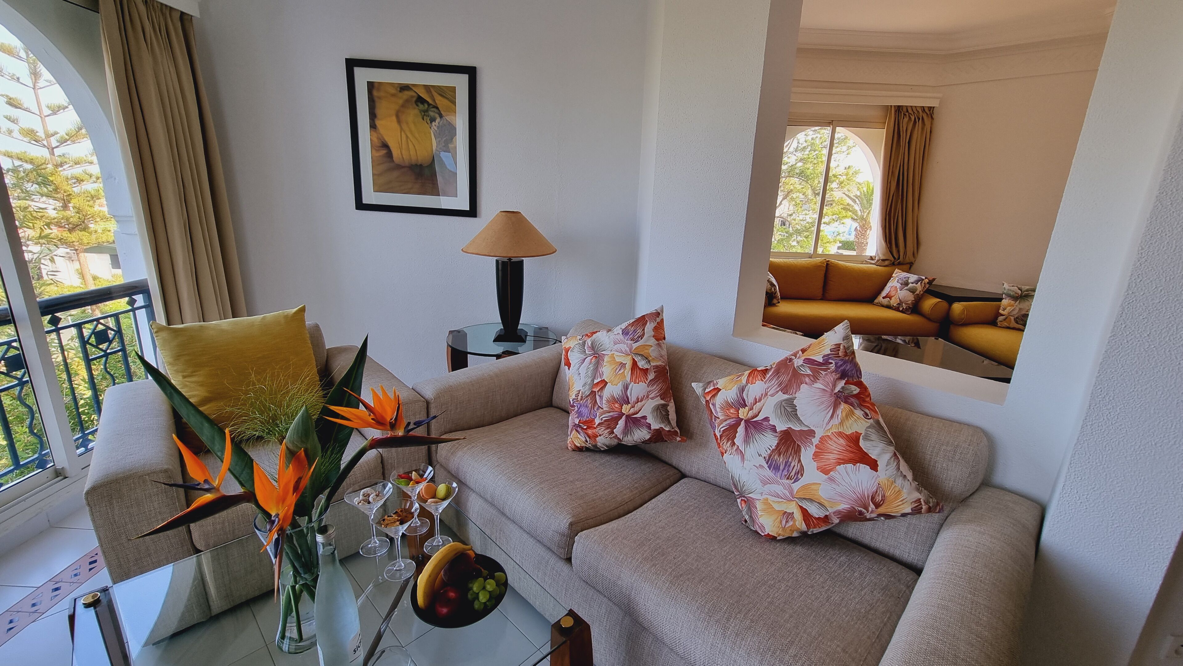 Salón con sofá, cojines decorativos, una mesa de centro de vidrio con bebidas y frutas, un jarrón con una flor de ave del paraíso, un cuadro en la pared y vistas al balcón.