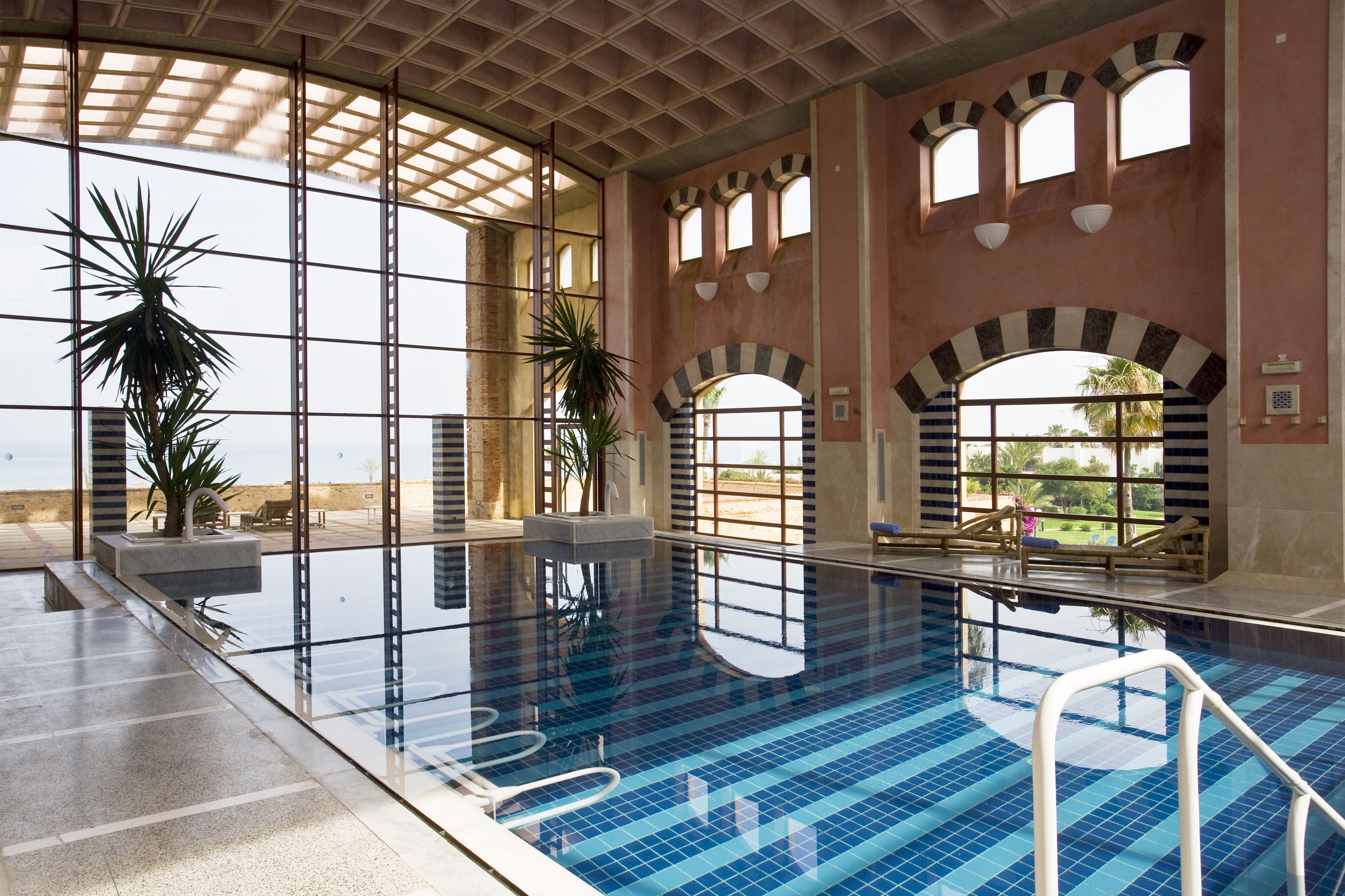 An indoor swimming pool with clear glass walls, arched windows, and potted plants.
