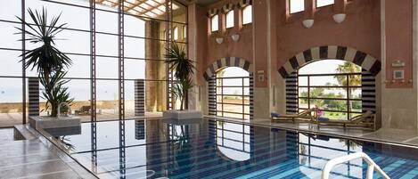 An indoor swimming pool with clear glass walls, arched windows, and potted plants.